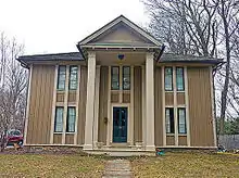 A two-story peaked-roofed wooden house with a projecting front full height porch topped by a pointed roof. It is sided with alternately thick and thin strips of wood running vertically.