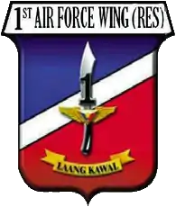 A shield-style crest primarily in red and blue, divided with a diagonal white bar.  The centre contains an symbolic military knife with the number '1' covering its blade.  The crest is headed by '1st Air Force Wing (Res)' in all-upper-case letters.  Below the knife is a yellow banner for its motto containing the phrase 'Laang Kawal'.