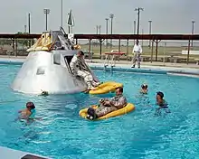 The Apollo spacecraft and orange rafts float in a pool, surrounded by divers