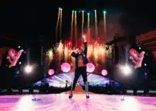 A man sings with a microphone as the stage behind him release numerous fireworks
