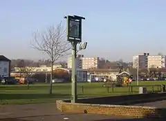 A photograph of an urban park with houses and flats in the distance. The park large, and mostly grass. A football goal is in the centre of the park, and a pub sign is in the foreground.