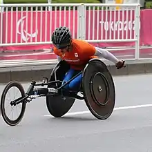 head down in orange shirt and racing chair with Tokyo 2020 on banners behind her