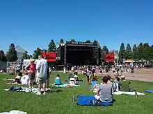 A bright blue sky over a lawn filled with people sitting on blankets or milling around, most facing a large stage with television screens on either side