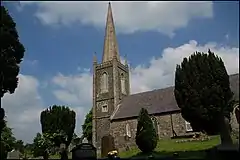 Photograph of a small stone church with tower and steeple, situated in a tidy graveyard.
