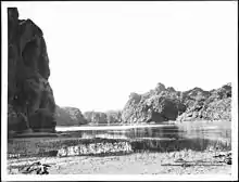 A steep rocky embankment looms in shadow at left. A tide mark is visible on the nearly vertical wall near the water line. The near bank is dried cracked mud. A few small pieces of driftwood sit on the bank. A rope lays on the bank at right. Across the placid river and in the distance rocky hills rise.