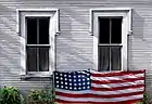 oil painting of a white clapboard house, two windows with flag draped along bottom sills, weeds along base