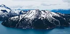 A lightly glaciated mountain rising above trees and a lake in the foreground.